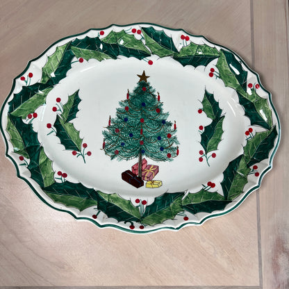 An oval white ceramic Christmas serving platter with a green holly and red berry border, featuring a decorated Christmas tree and wrapped gifts in the center, shown on a light wood surface.