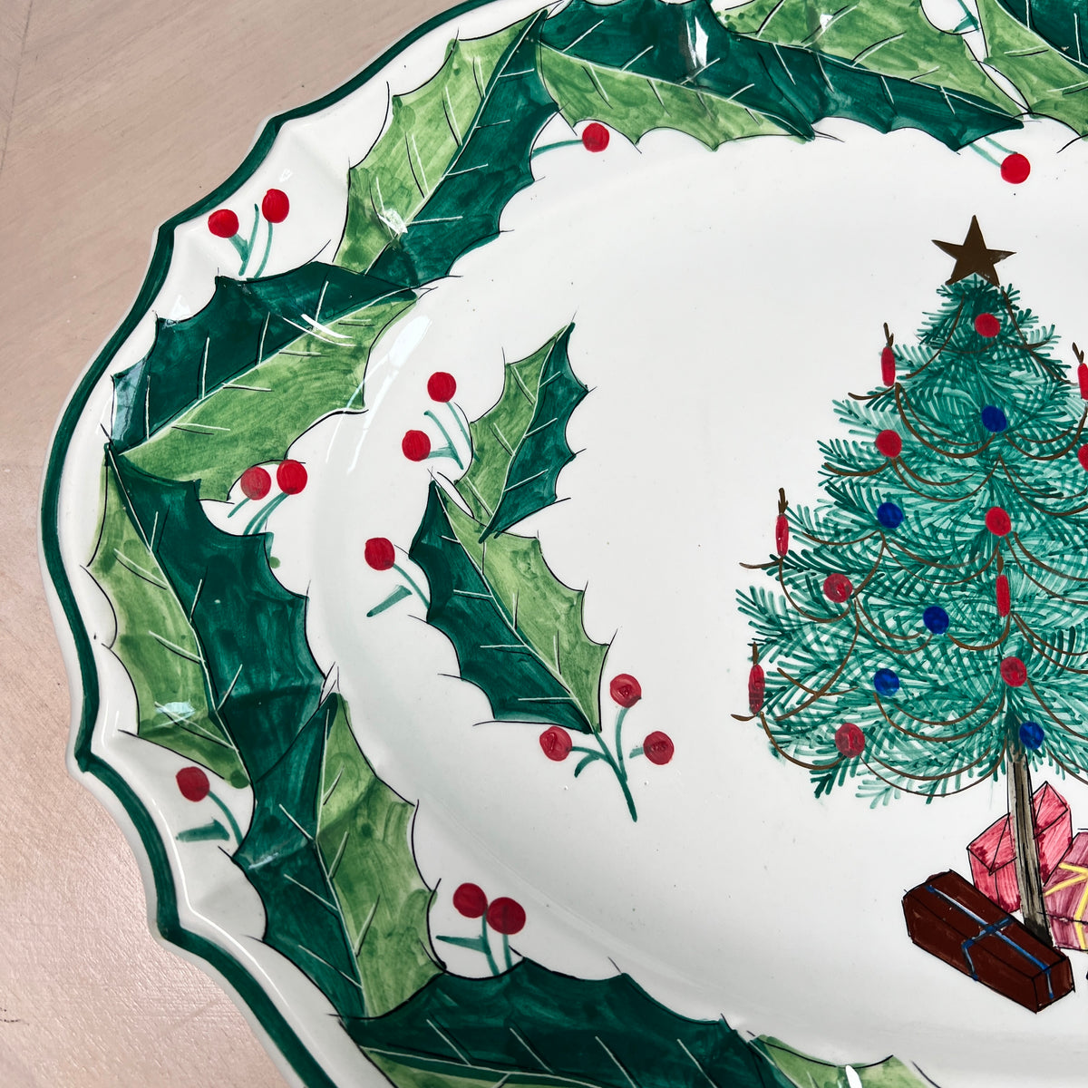 A close-up of a white ceramic Christmas plate with a green holly border, red berries, and a decorated Christmas tree illustration on the right side.