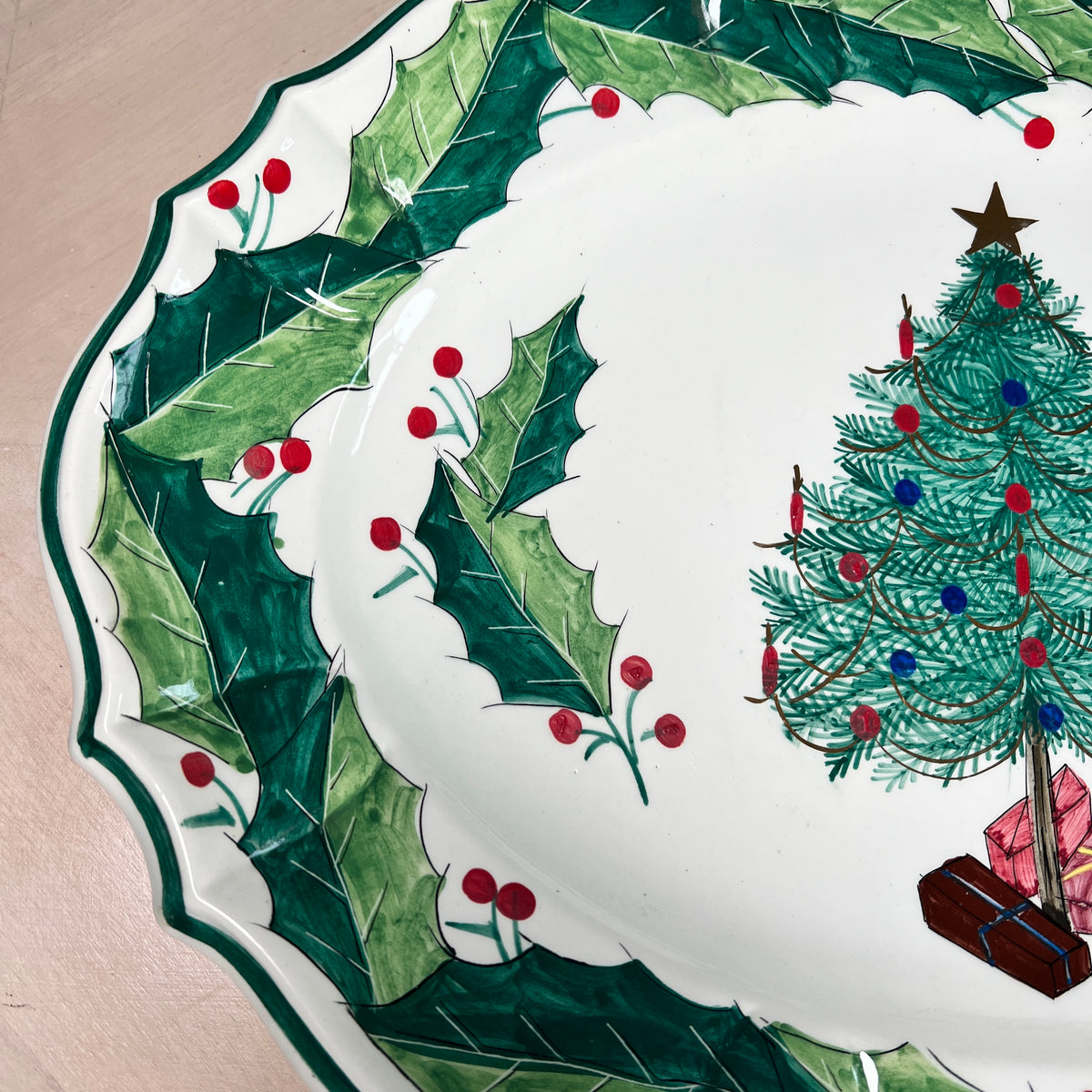 A close-up of a white ceramic plate with a scalloped edge, decorated with a green holly wreath and a Christmas tree illustration on the right side.