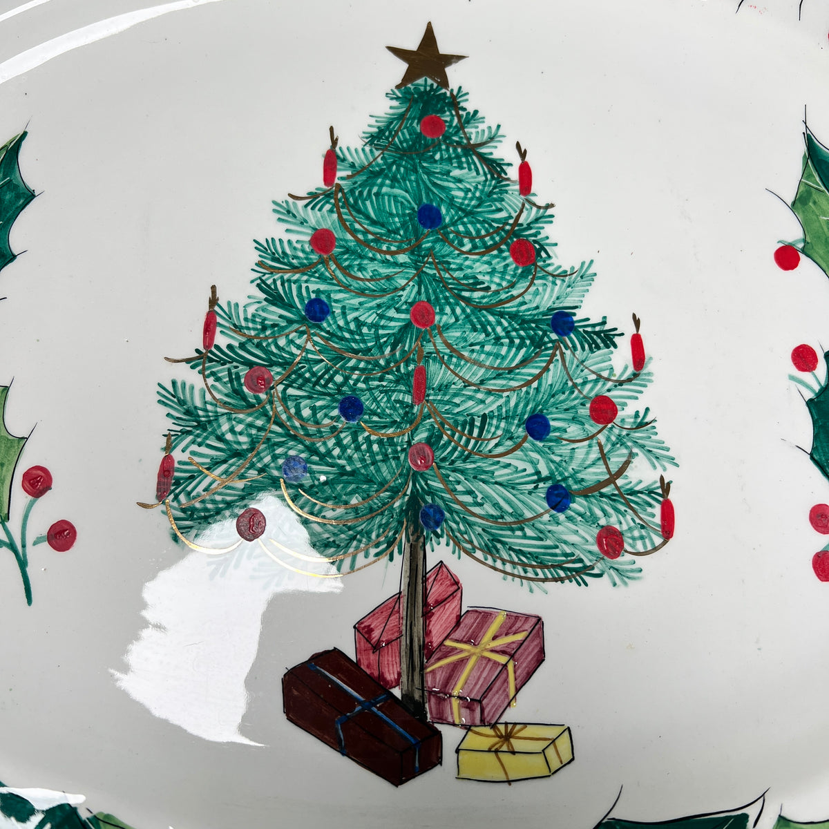 A close-up of a white ceramic plate or dish featuring a hand-painted Christmas tree illustration with red and blue ornaments, wrapped in gold ribbon, a gold star topper, and three presents underneath.