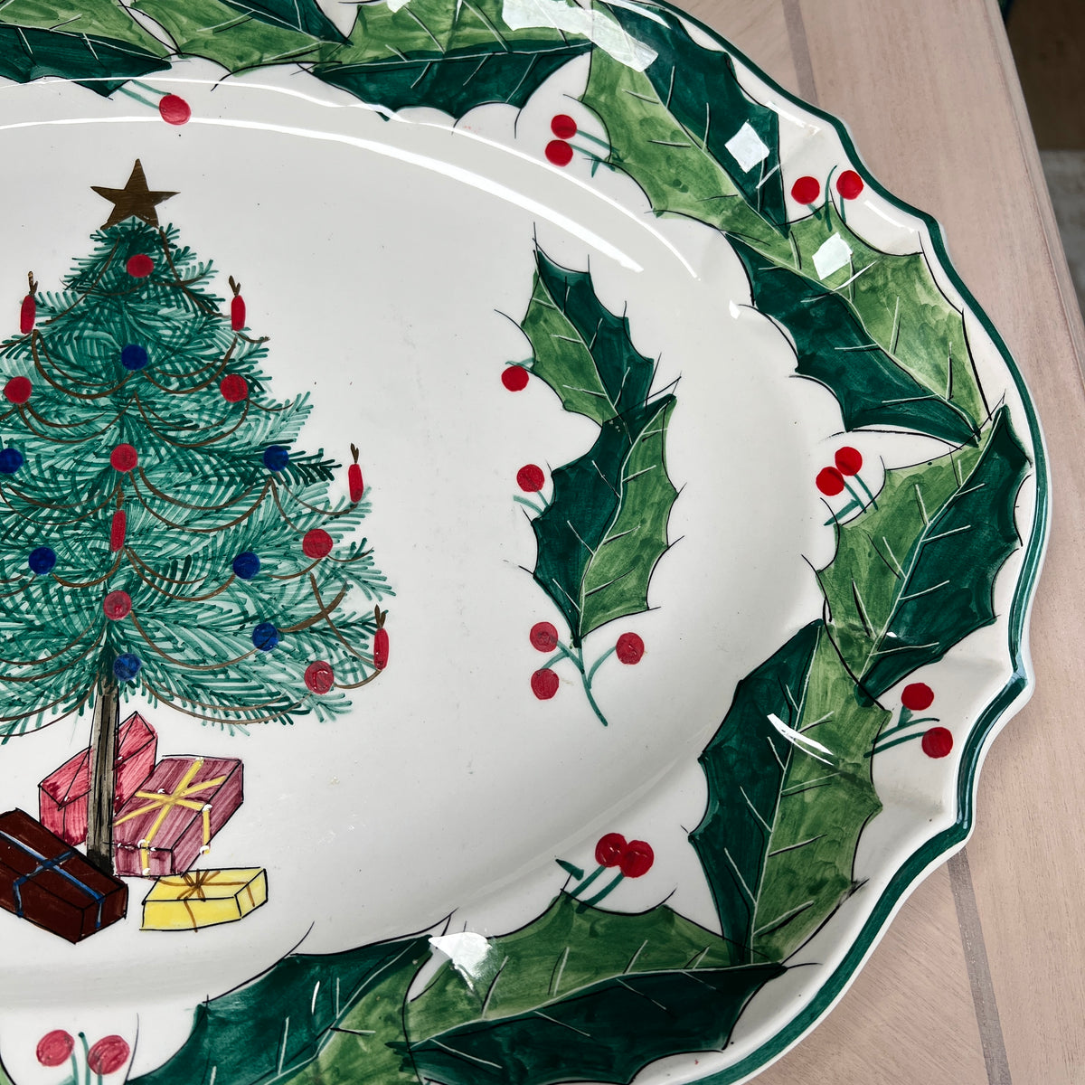A white ceramic Christmas plate with a green holly wreath border, red berries, a decorated Christmas tree, and wrapped gifts, photographed on a light wood surface.