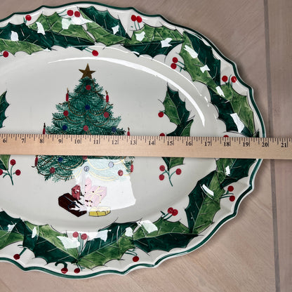 A white ceramic Christmas platter with a green holly and red berry border, featuring a painted Christmas tree and Santa and elf scene. A wooden ruler is placed across the center to show the diameter.