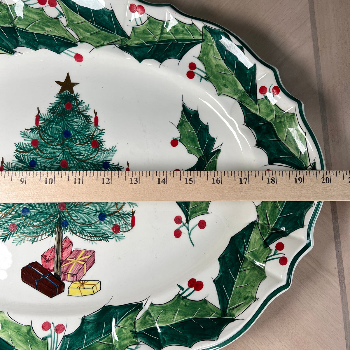 A white ceramic Christmas-themed serving platter with a green holly and berry border, a decorated Christmas tree, and wrapped gifts. A wooden ruler is placed horizontally across the center to show the platter's width.