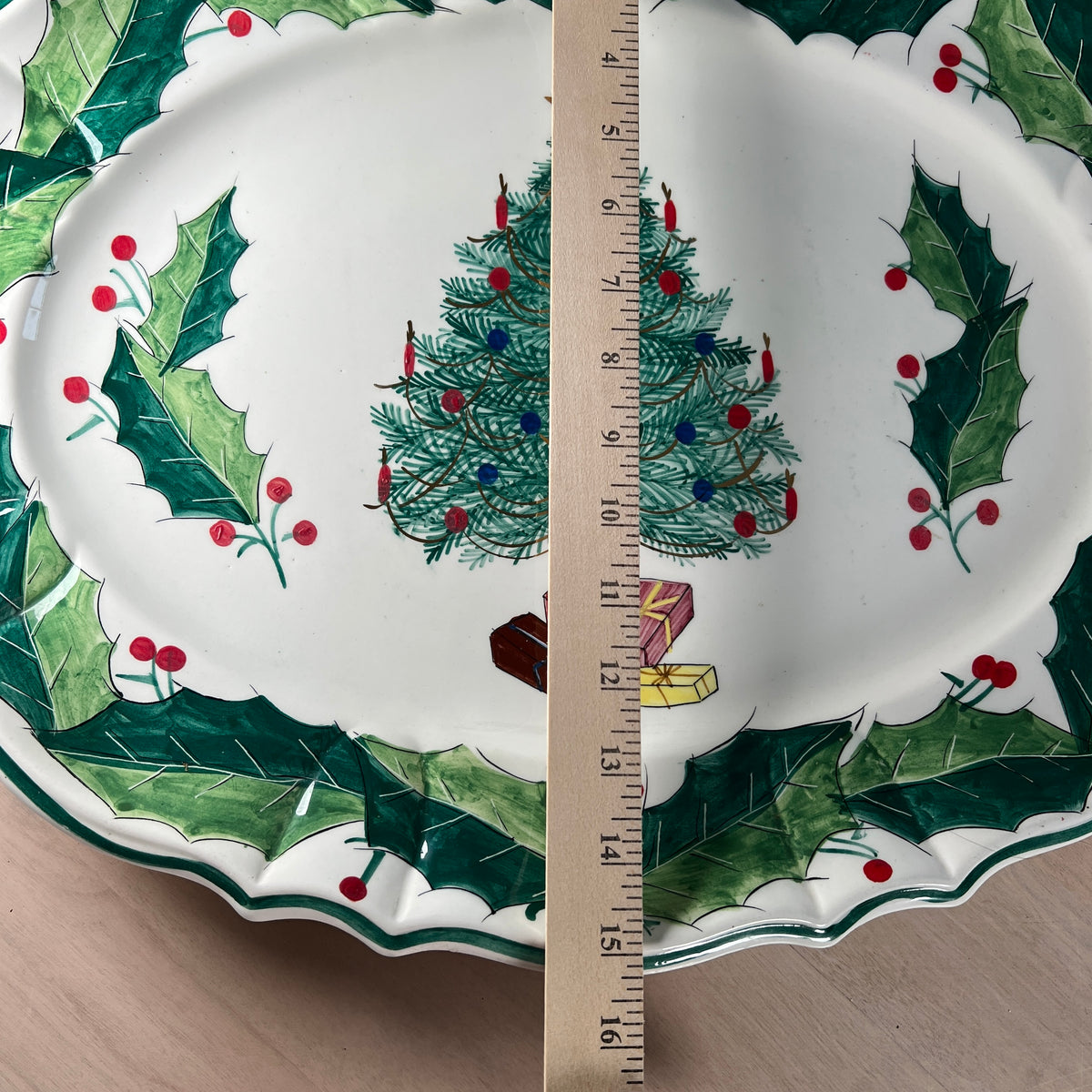 A white ceramic Christmas plate with a green holly and berry border and a decorated Christmas tree in the center, shown with a wooden ruler for scale on a light wood surface.