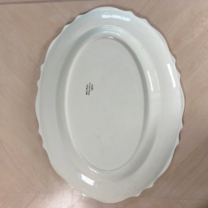 An oval white ceramic serving platter with a scalloped rim, photographed from directly above on a light wood floor.
