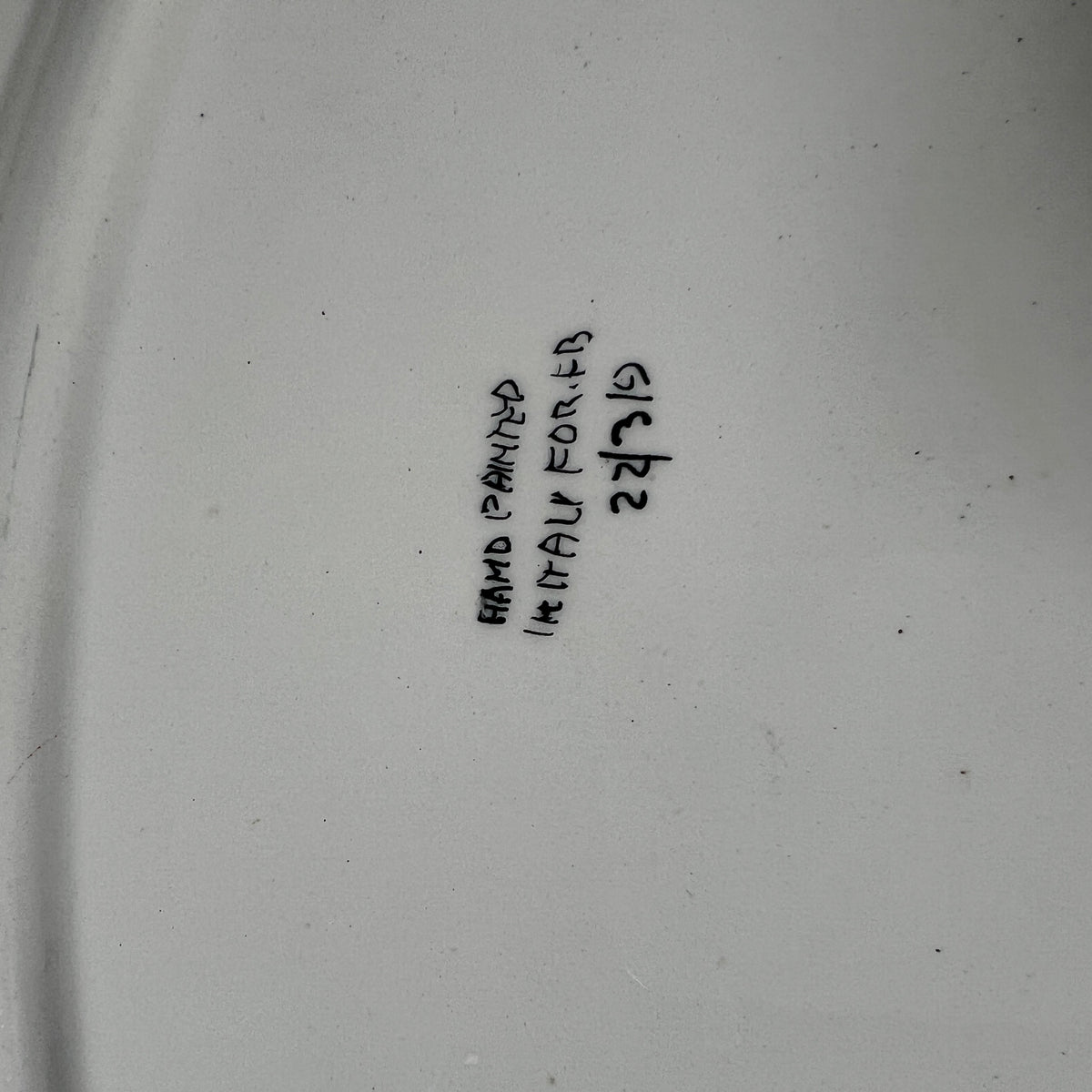 A close-up of a white ceramic surface with handwritten black text reading 'HAND PAINTED' and 'Italy FOR FB' along with a handwritten 22/319