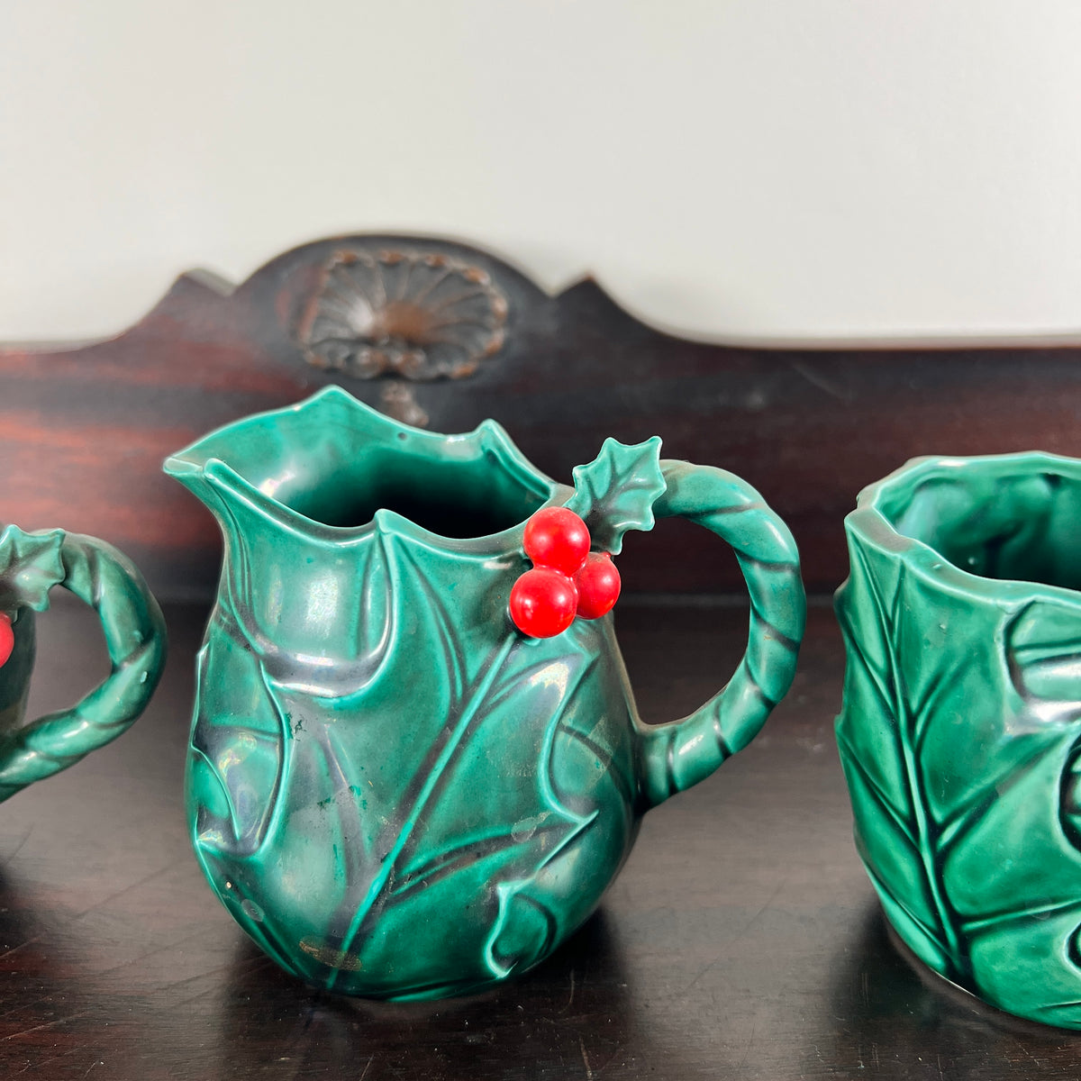 A green ceramic pitcher shaped like a holly leaf, decorated with three red berries, shown on a dark wooden table next to matching green ceramic cups.