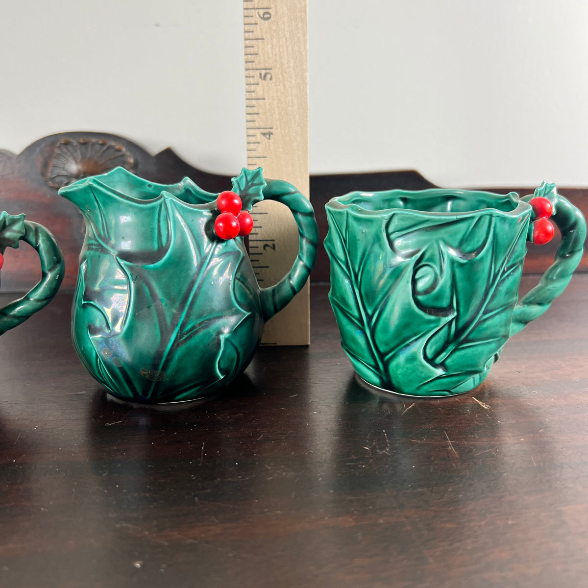 A pair of green ceramic Christmas cups with embossed holly leaves and red berries, shown on a dark wooden table with a wooden ruler standing behind them for scale.