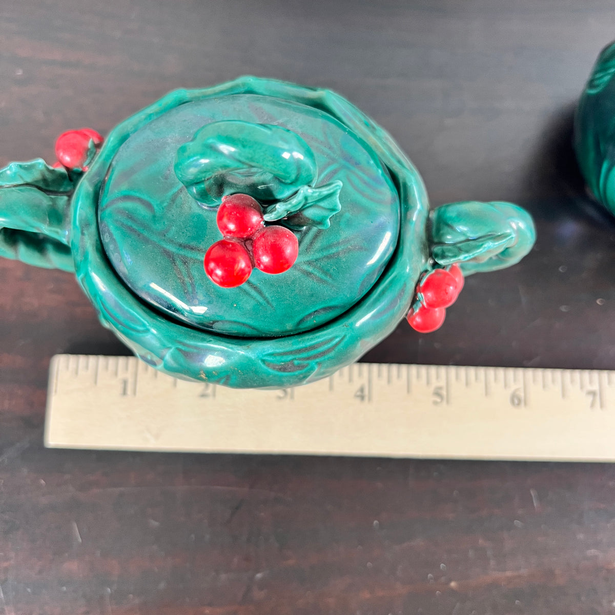 A green ceramic teapot with a lid, decorated with red berries and green leaves, shown next to a wooden ruler on a dark wooden surface.