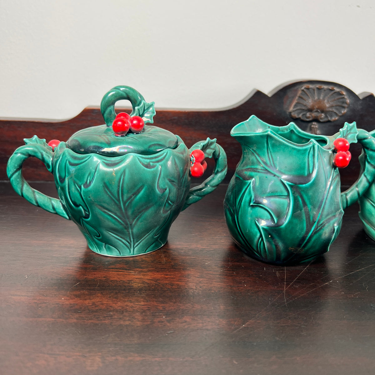 A green ceramic teapot and matching pitcher set decorated with embossed holly leaves and red berry accents, displayed on a dark wooden table.