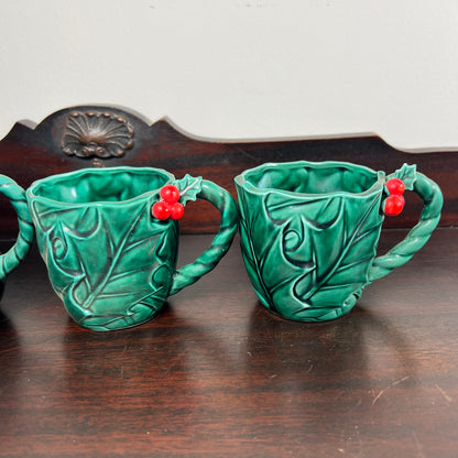Two green ceramic mugs shaped like leaves with red holly berries, displayed on a dark wooden surface.