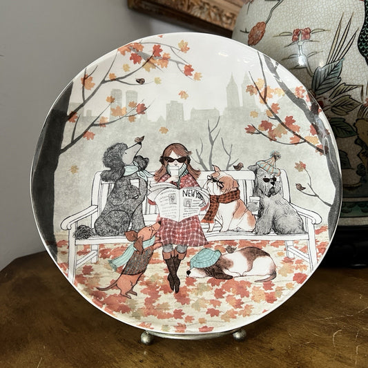 Decorative plate with a design of a woman reading a newspaper surrounded by dogs on a bench, with autumn leaves and cityscape in the background.