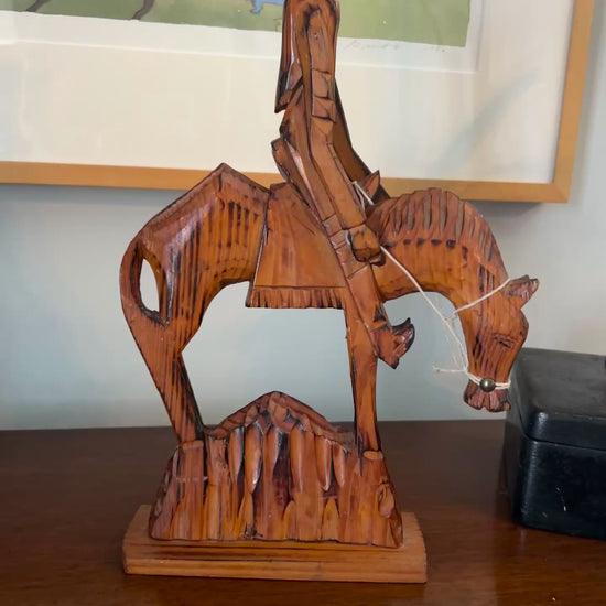 Video of Wooden sculpture of a cowboy on horseback on a table with a painting and plant in the background.