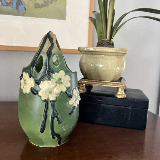 Green ceramic vase with floral design on a wooden surface, next to a potted plant.