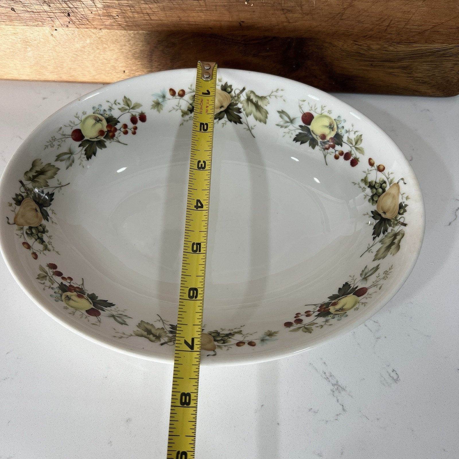 Decorative ceramic bowl with fruit pattern on a white surface, measured by a tape measure.