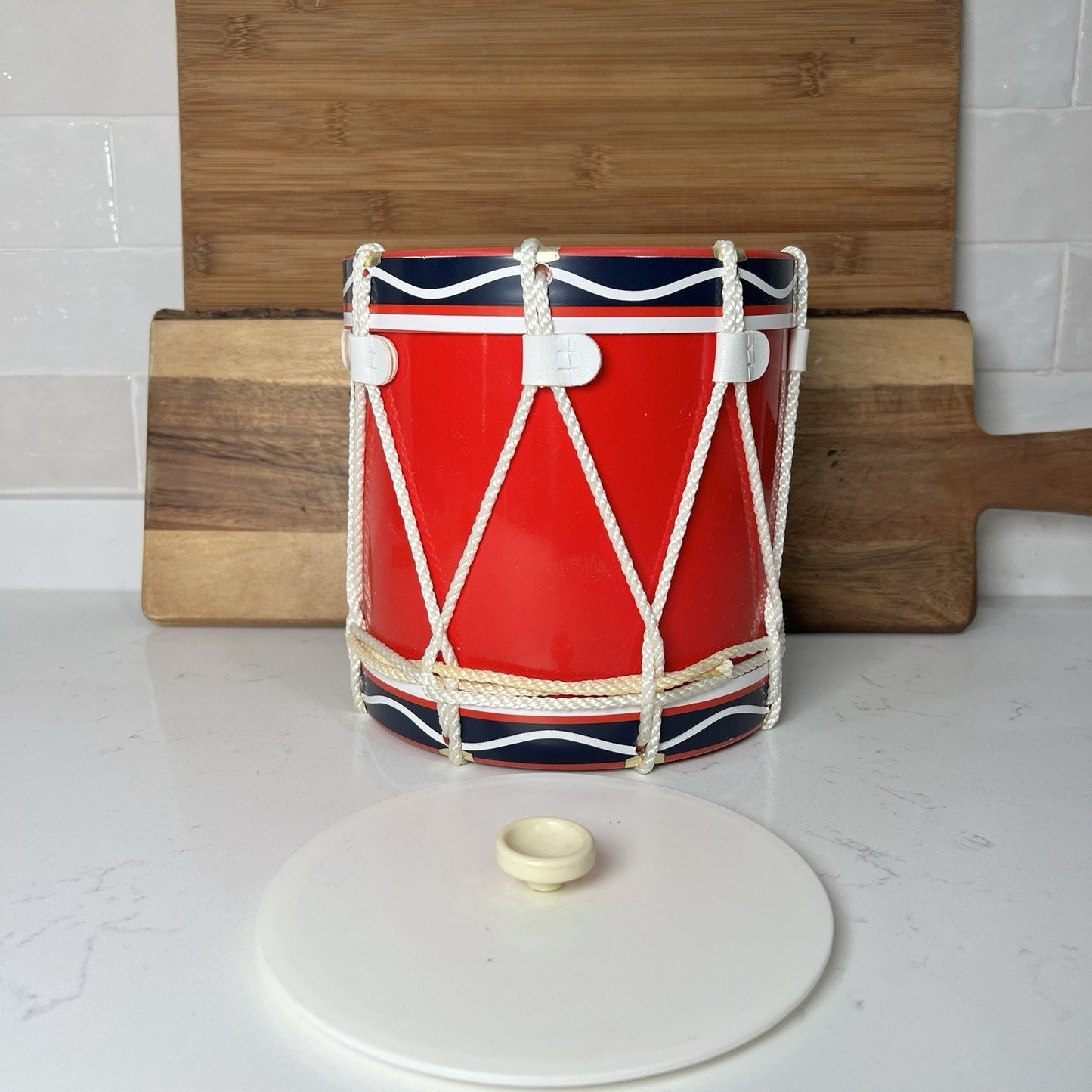 Red drum-shaped container with rope detailing on a white surface with wooden cutting boards in the background.