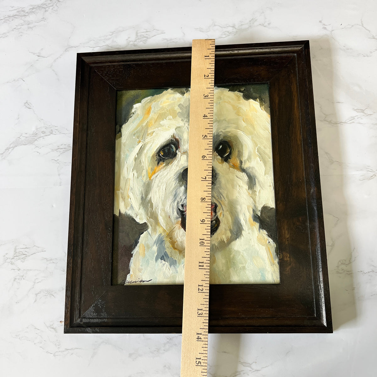 A framed portrait painting of a white dog, shown with a wooden ruler standing vertically to indicate the size of the dark brown frame and artwork against a light marble background.