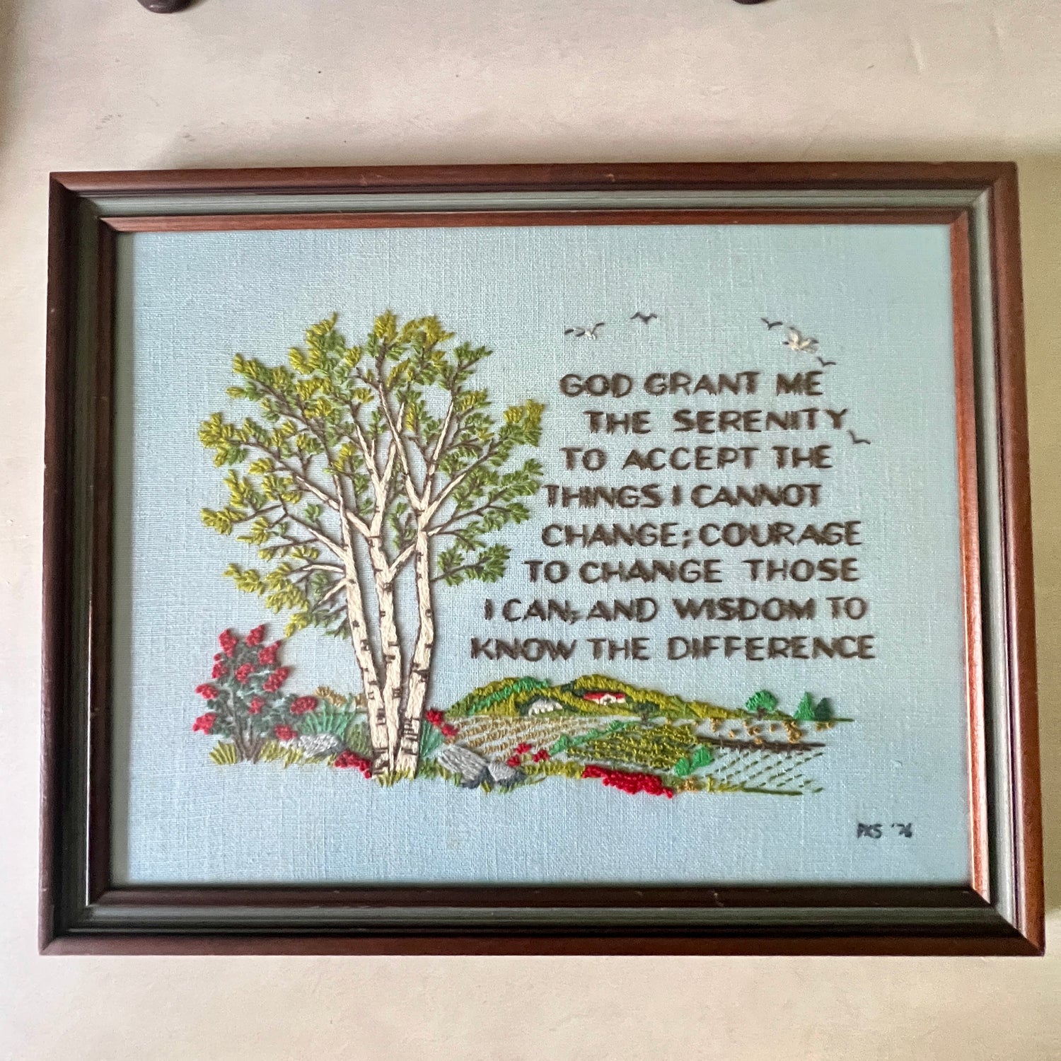 A framed embroidered wall hanging with a light blue background, featuring a birch tree, small birds, red flowers, and a landscape with the Serenity Prayer text: 'God grant me the serenity to accept the things I cannot change; courage to change those I can, and wisdom to know the difference.' The piece is enclosed in a dark brown wooden frame and photographed against a beige wall.