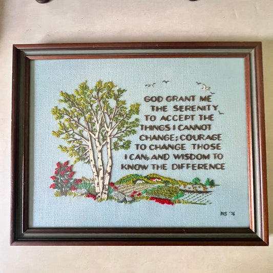 A framed embroidered wall hanging with a light blue background, featuring a birch tree, small birds, red flowers, and a landscape with the Serenity Prayer text: 'God grant me the serenity to accept the things I cannot change; courage to change those I can, and wisdom to know the difference.' The piece is enclosed in a dark brown wooden frame and photographed against a beige wall.