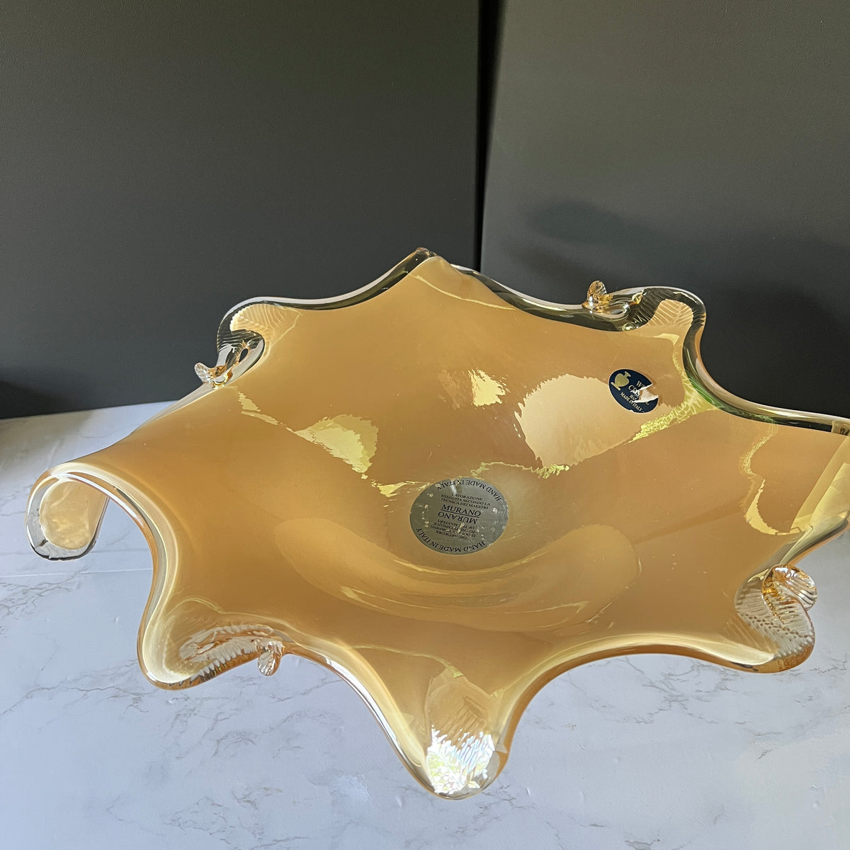 A translucent amber-gold glass bowl with a wavy, scalloped rim and small integrated handles, photographed on a light marble surface against a dark gray background.