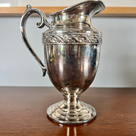 A small vintage silverplate creamer pitcher with a curved spout and ornate handle sits on a wooden table. The body is rounded with a pedestal base, and a decorative floral band wraps around the upper section. The surface shows tarnish and age patina.